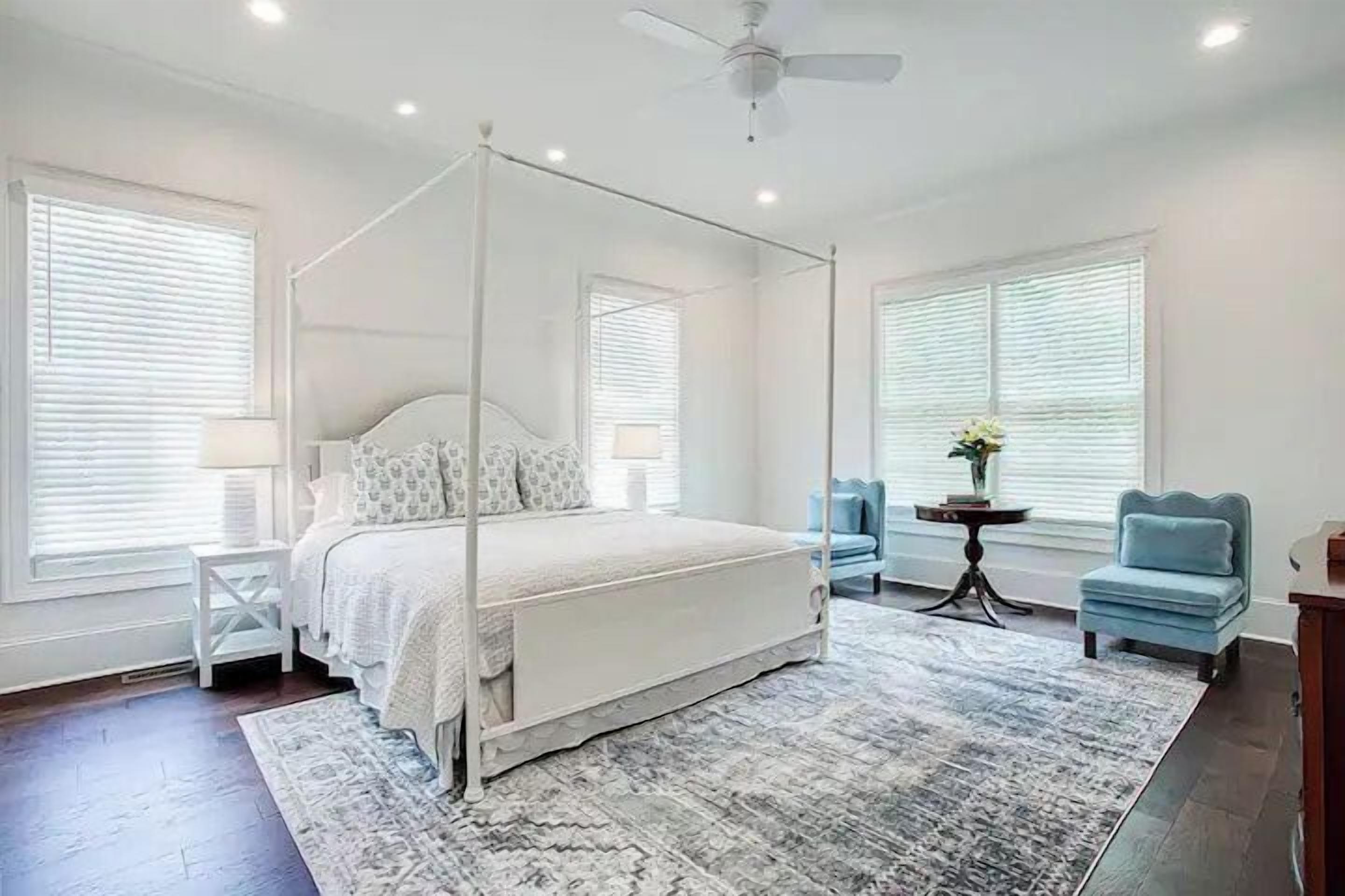 A four-poster bed with a canopy, bedside tables, a small round table with a vase, two armchairs, and a ceiling fan in a bedroom.