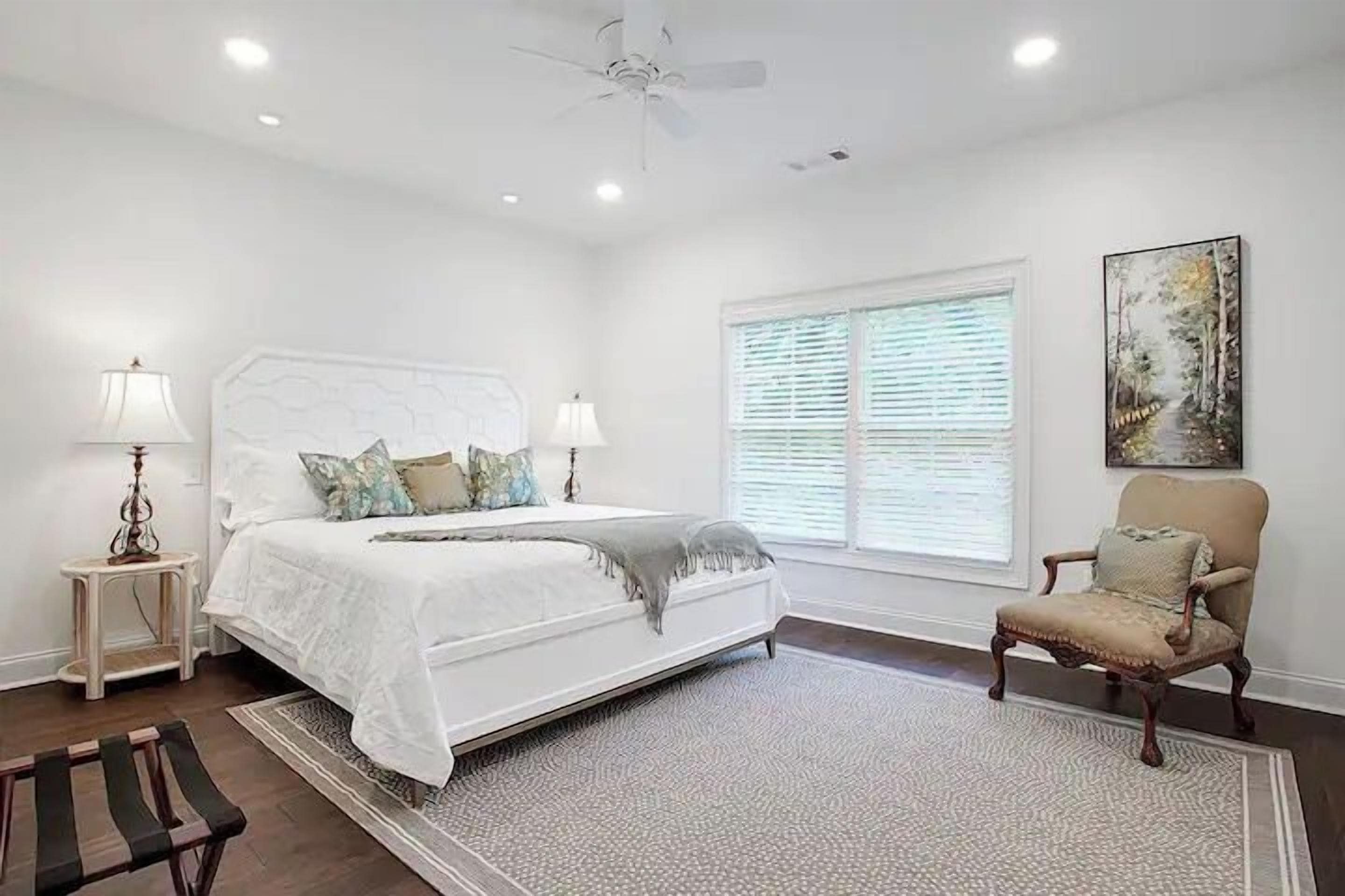 A bedroom with a large bed, bedside lamps, a chair, a window with blinds, and a ceiling fan.