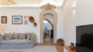 A living room with a grey sofa, a TV, and a decorative archway leading to a dining area. - Casa Cordovil Evora (Évora)