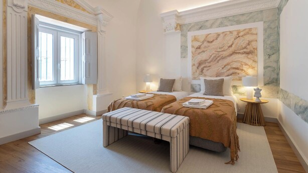 A bedroom with a large bed, two bedside tables with lamps, a bench, and a marble wall. - Casa Cordovil Evora (Évora)