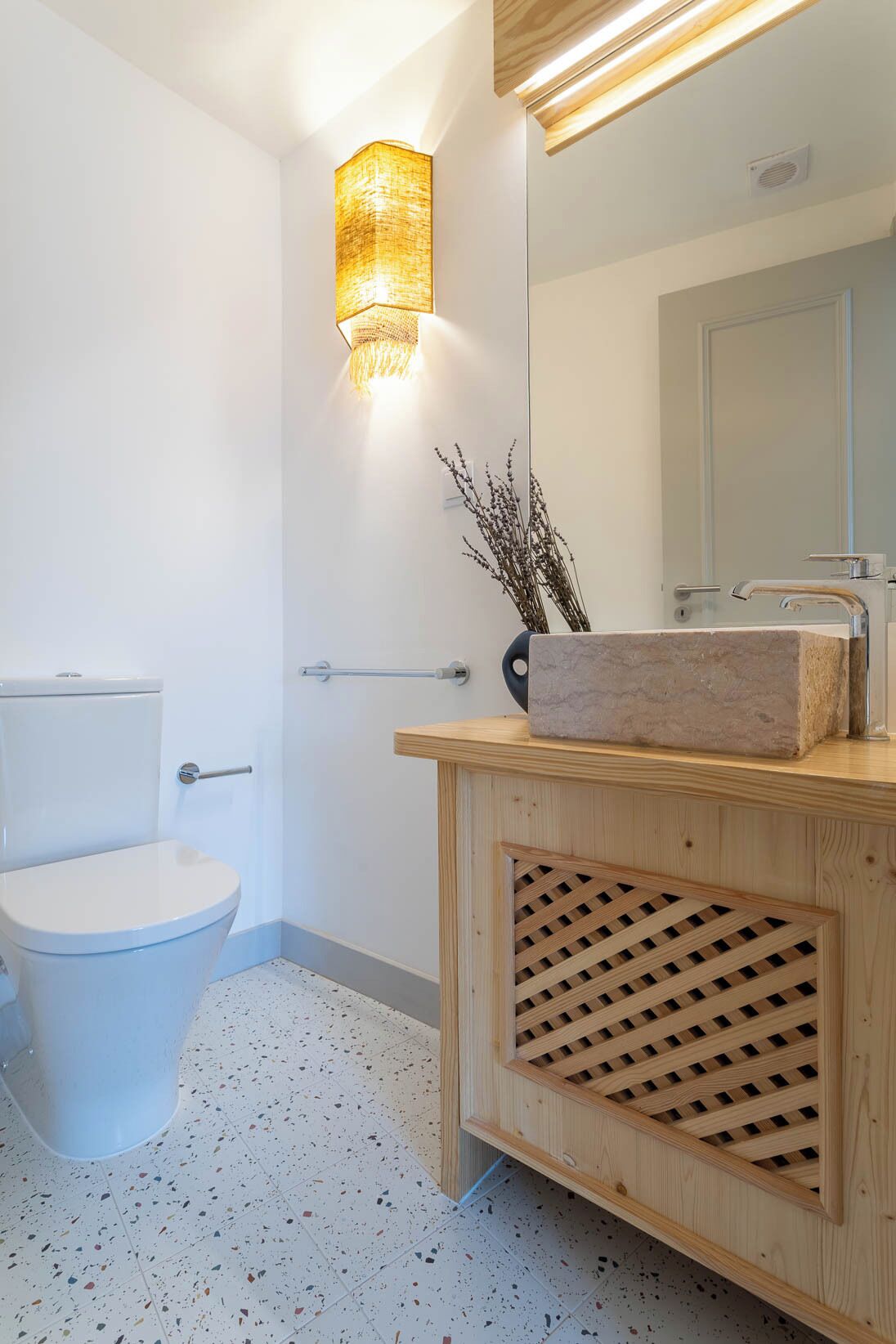 Un bagno moderno con un WC, un mobiletto in legno con lavabo in marmo, un lampadario a parete e uno specchio.