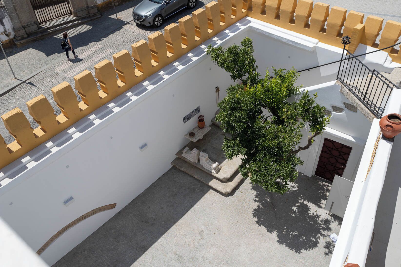 An aerial view of a courtyard with a tree, a small staircase, and a white building with yellow railings.