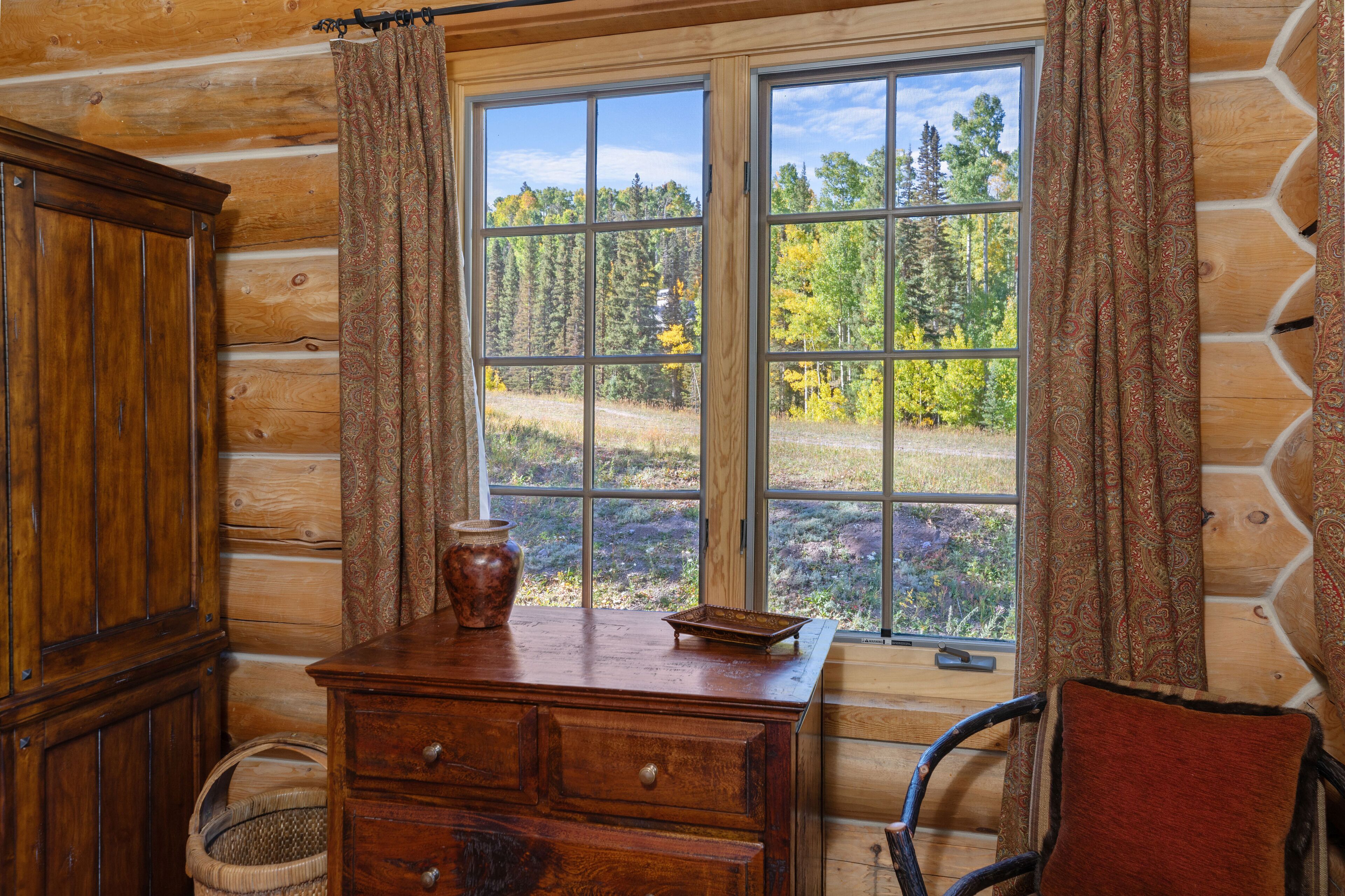 Une chambre aménagée dans un chalet en rondins, avec une grande fenêtre, une commode en bois, un fauteuil en osier et un panier.