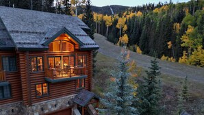 A log cabin with a stone foundation, a balcony with a railing, and a view of a forested hillside. - First Tracks on Sundance - Slope-side Skiing! Views - Private Hot Tub - A/C ! (Mountain Village)