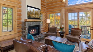 A log cabin interior with a stone fireplace, leather furniture, and large windows offering views of trees and a mountain. - First Tracks on Sundance - Slope-side Skiing! Views - Private Hot Tub - A/C ! (Mountain Village)