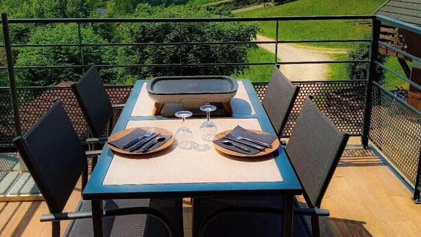 Un espace de restauration en plein air avec une table dressée pour deux personnes, offrant une vue panoramique sur un paysage pittoresque composé de collines et d’une étendue d’eau.