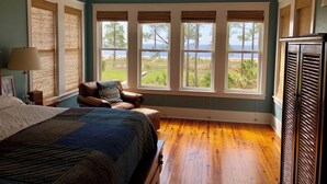 A bedroom with a large bed, a chair, and a view of the beach through multiple windows.
