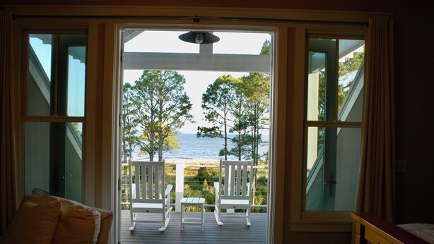 A room with a large window overlooking a deck with white chairs and a view of the sea.