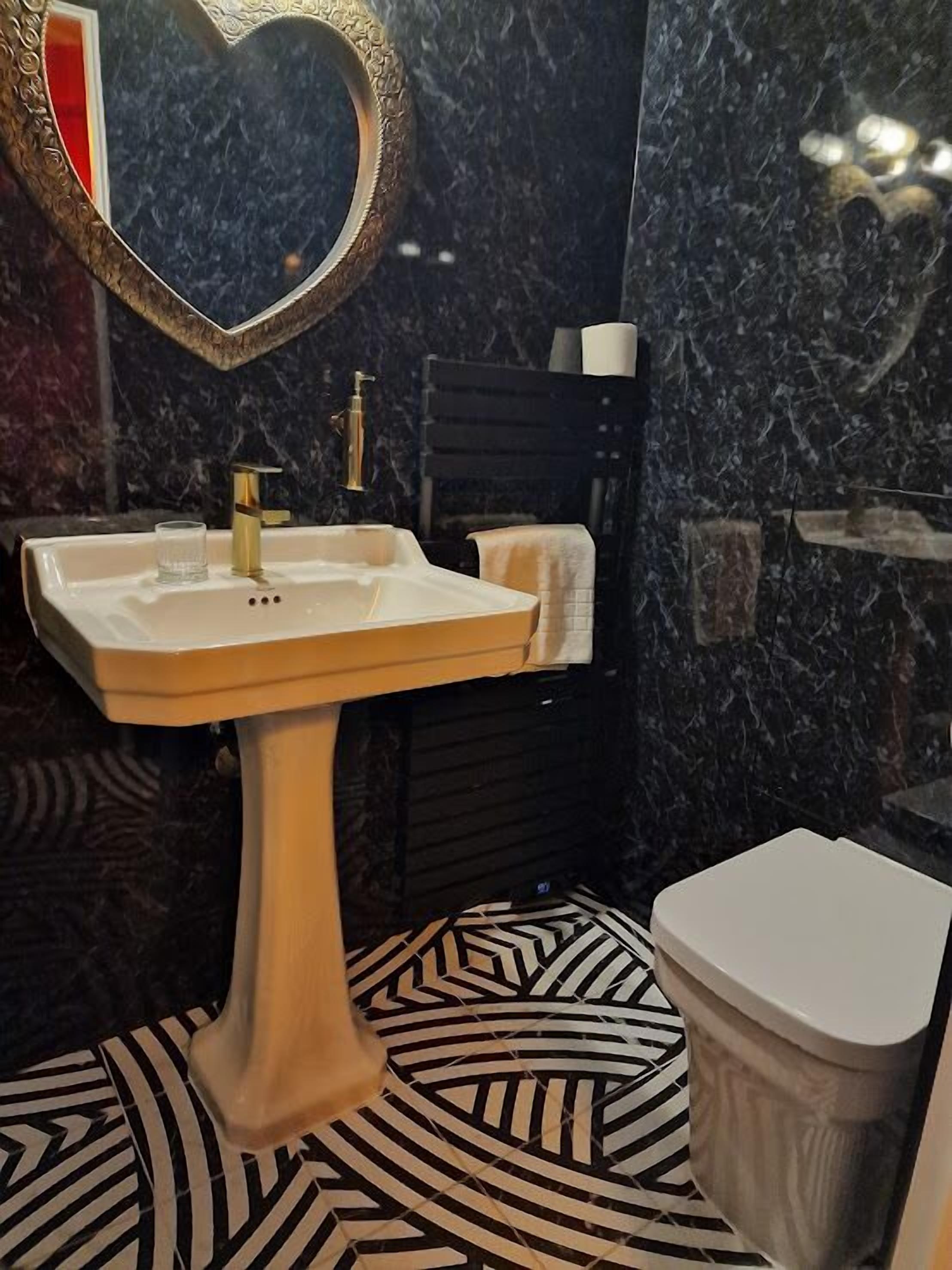 A bathroom with a white pedestal sink, a heart-shaped mirror, a black towel, and a patterned floor.