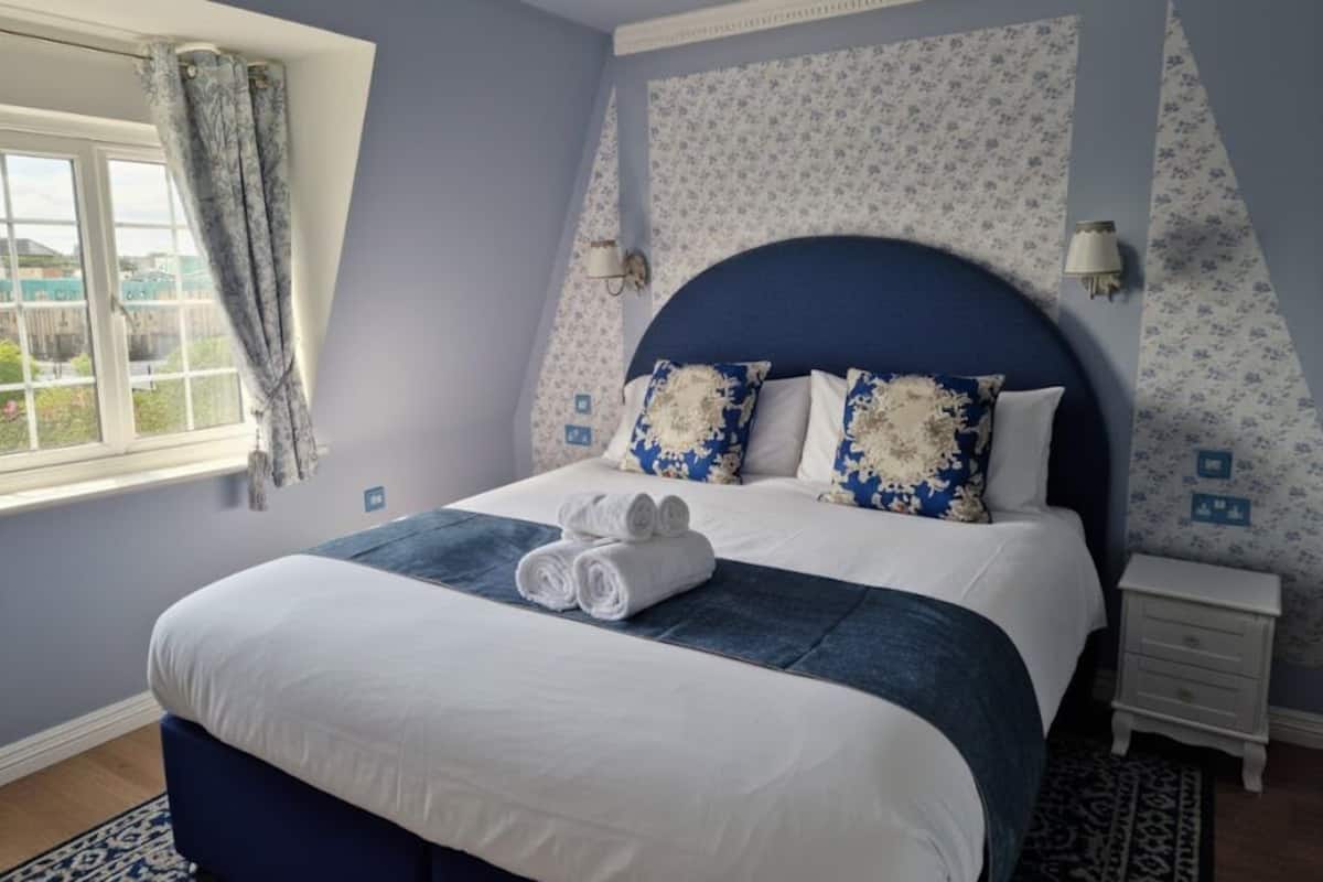 A neatly made bed with a blue headboard, white bedding, and decorative pillows. There is a window with curtains, a bedside table, and a patterned rug.