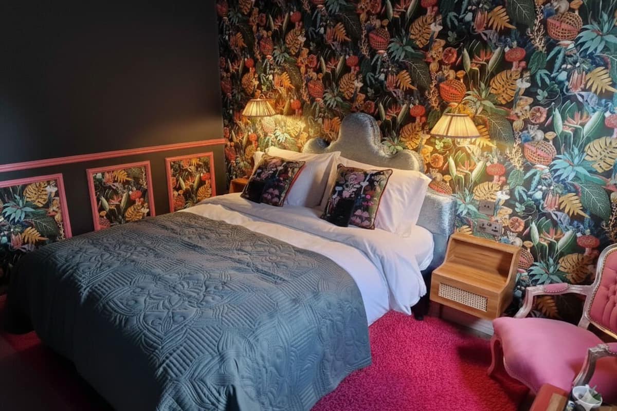 A bedroom with a bed, a nightstand, a chair, and a wall with a tropical floral pattern.