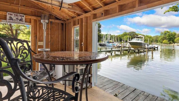 A wooden deck with a round table and chairs, overlooking a calm lake with boats.