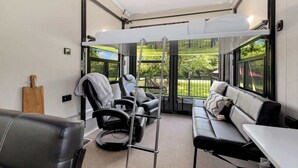 A modern room with a sofa, two massage chairs, a table, and a view of greenery through large windows.