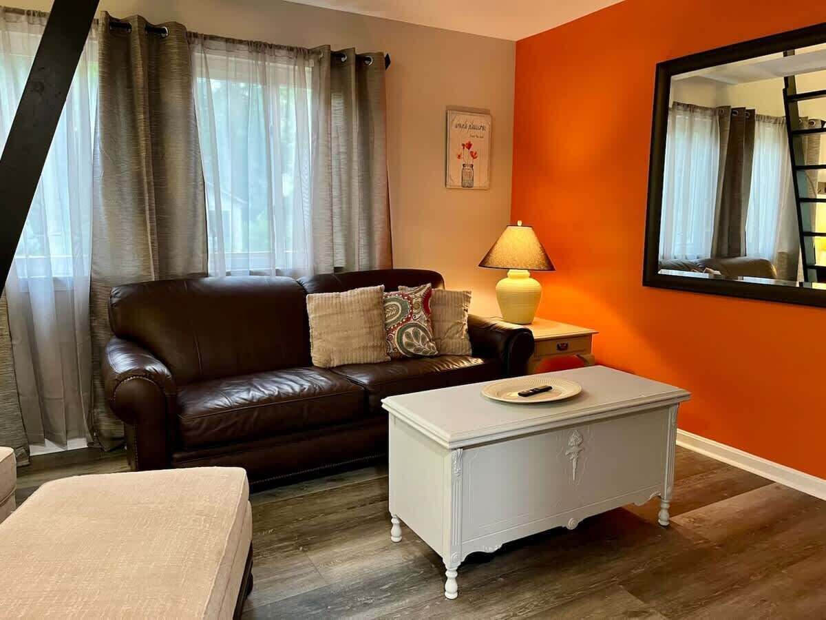A living room with a brown leather sofa, a white side table with a lamp, and a large mirror on the orange wall.