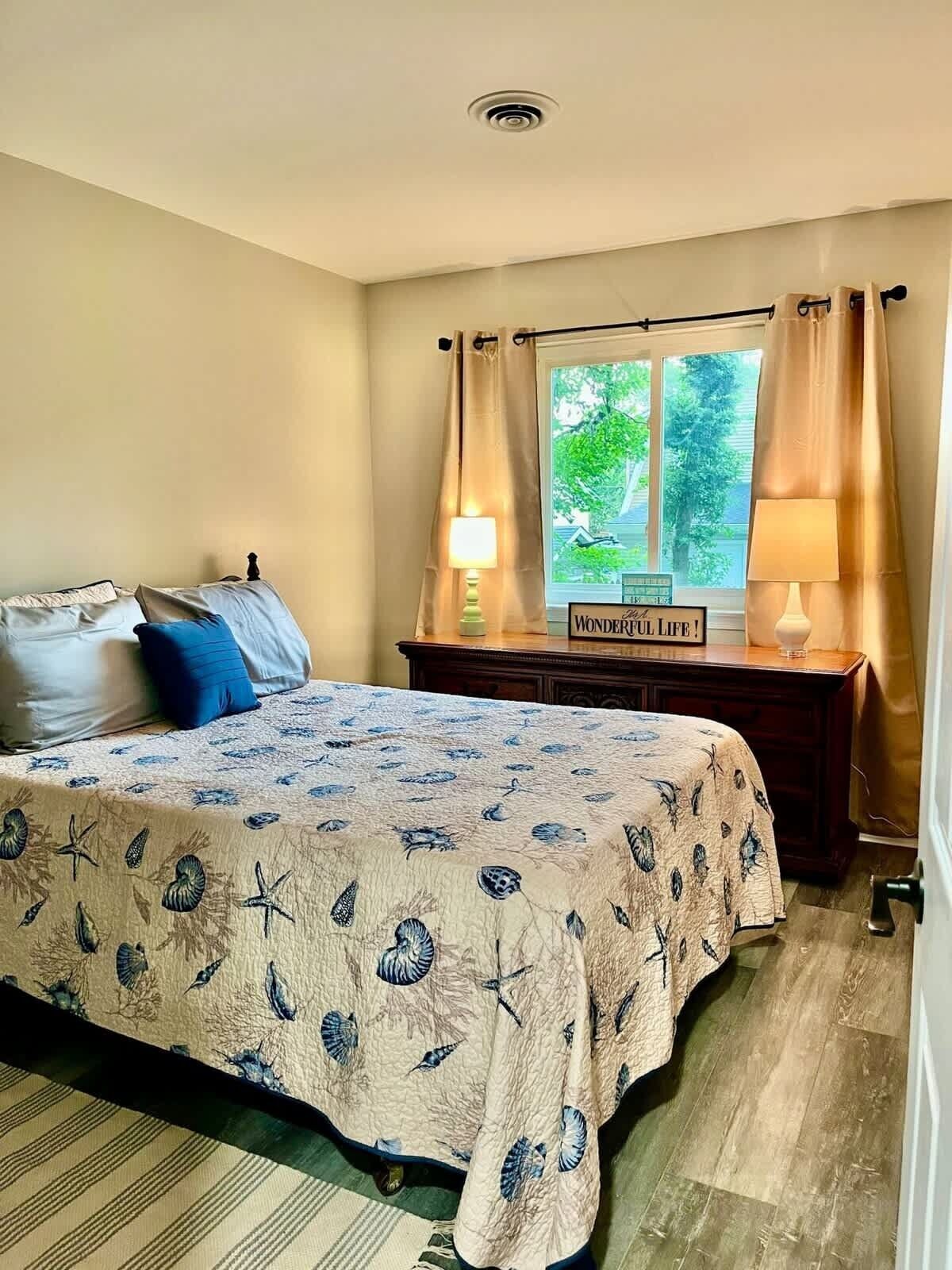 A bedroom with a bed, a nightstand, two lamps, a window with curtains, and a sign on the window.