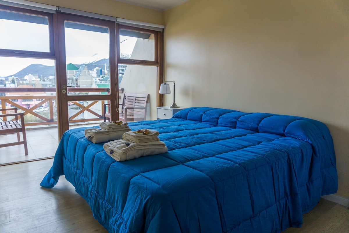Ein Zimmer mit einem großen Bett, einem Sessel, einem Nachttisch und einem Fenster mit Blick auf Gebäude und Berge.