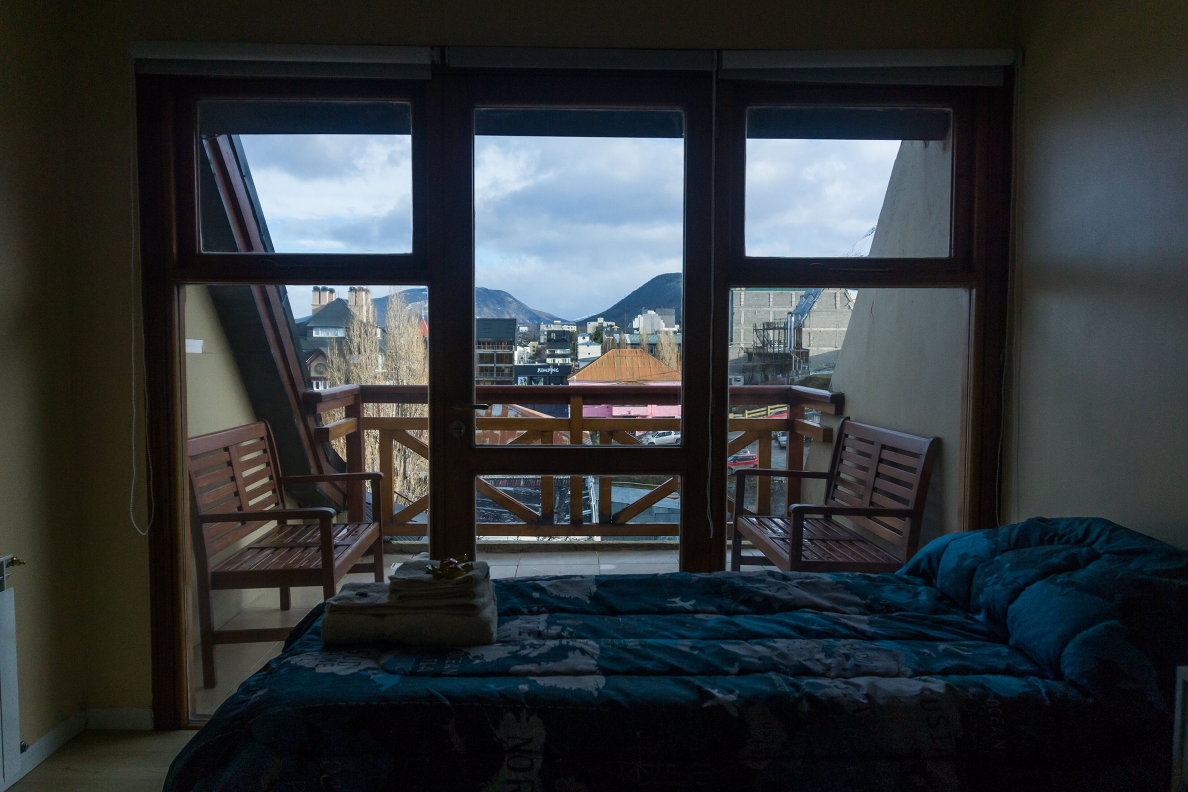 A bedroom with a large window, a bed, and a view of a balcony with chairs and a table.