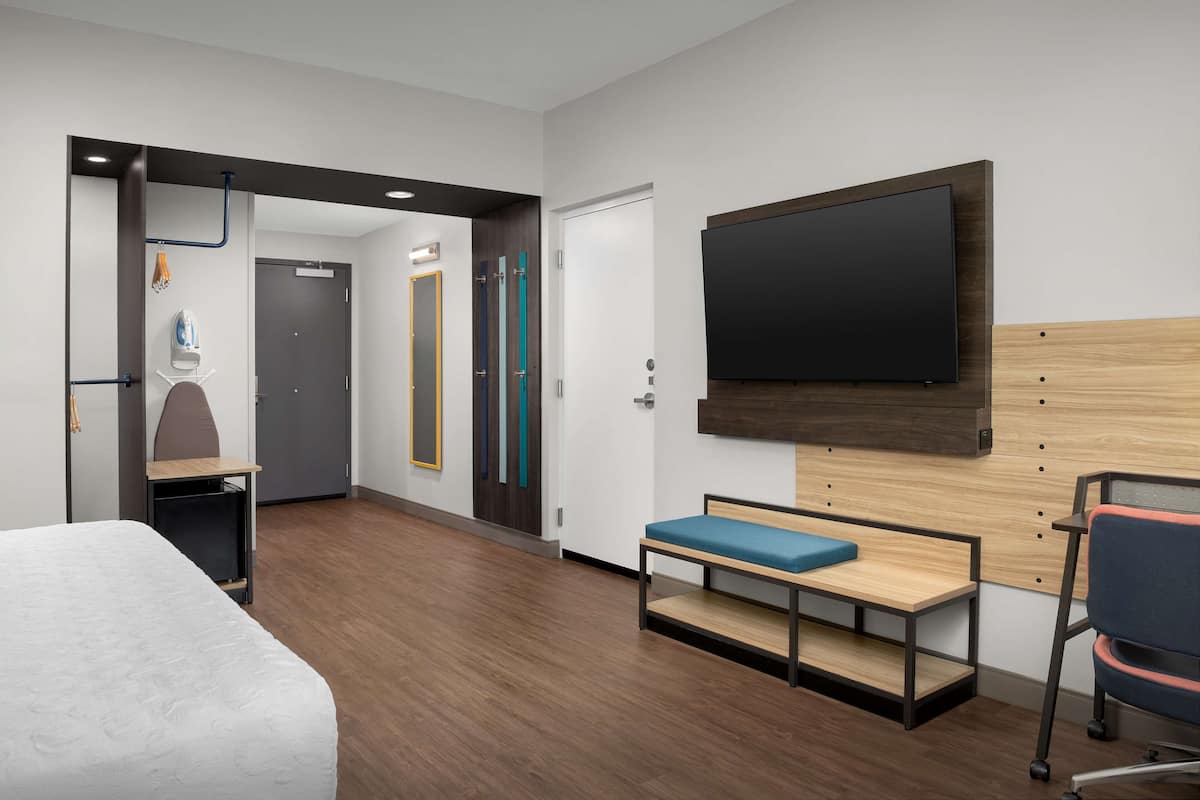 Une chambre d’hôtel comprenant un lit, un banc, un téléviseur fixé au mur, une porte avec un miroir et un porte-serviettes.