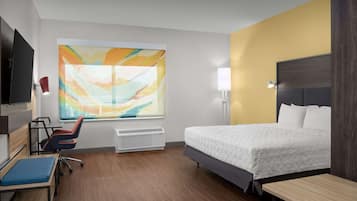 Quarto de hotel moderno com uma cama grande, uma escrivaninha, uma cadeira, uma TV e um quadro abstrato grande na parede.