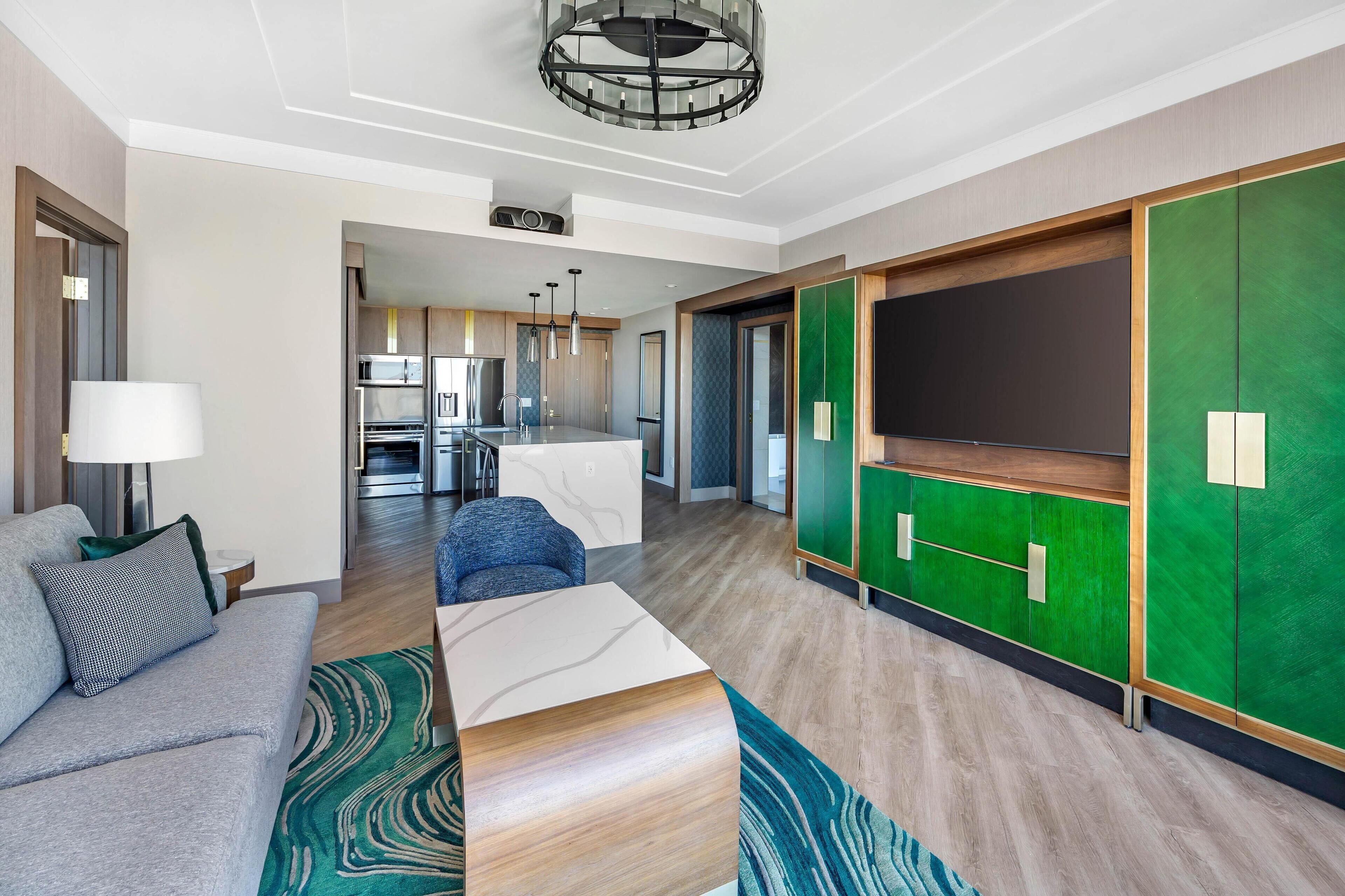 A modern hotel room with a sofa, a coffee table, a green accent wall, and a view into the kitchen area.