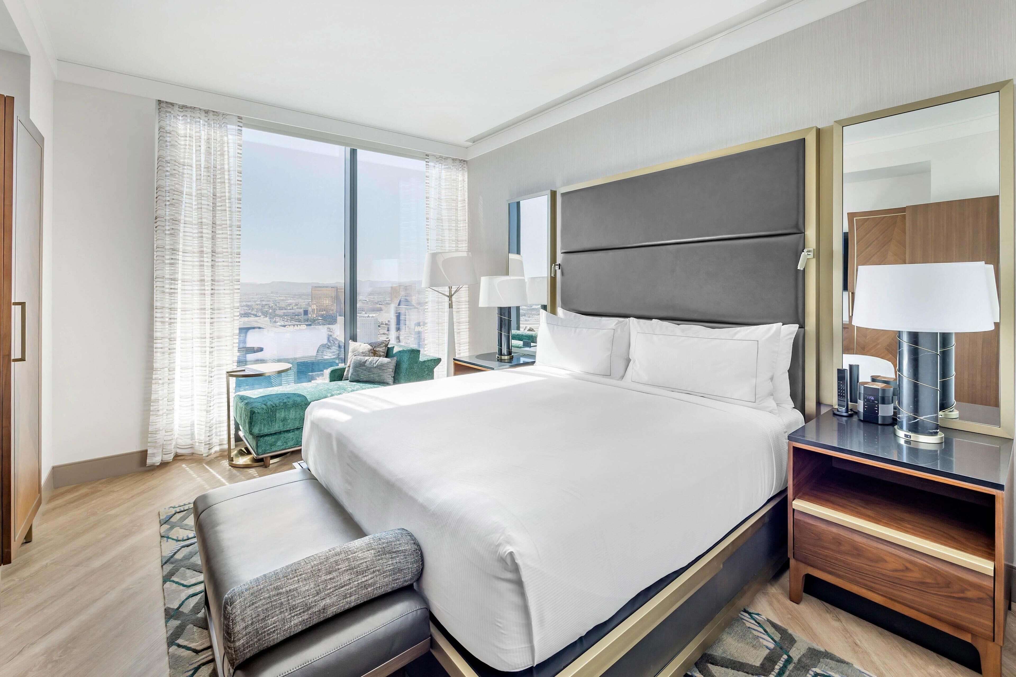 A modern hotel room with a large bed, a seating area with a green sofa, a nightstand with a lamp, and a view of the city through large windows.
