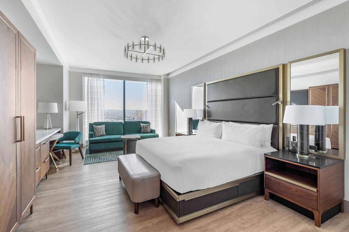 A modern hotel room with a large bed, a desk, a sofa, and a view of the city.