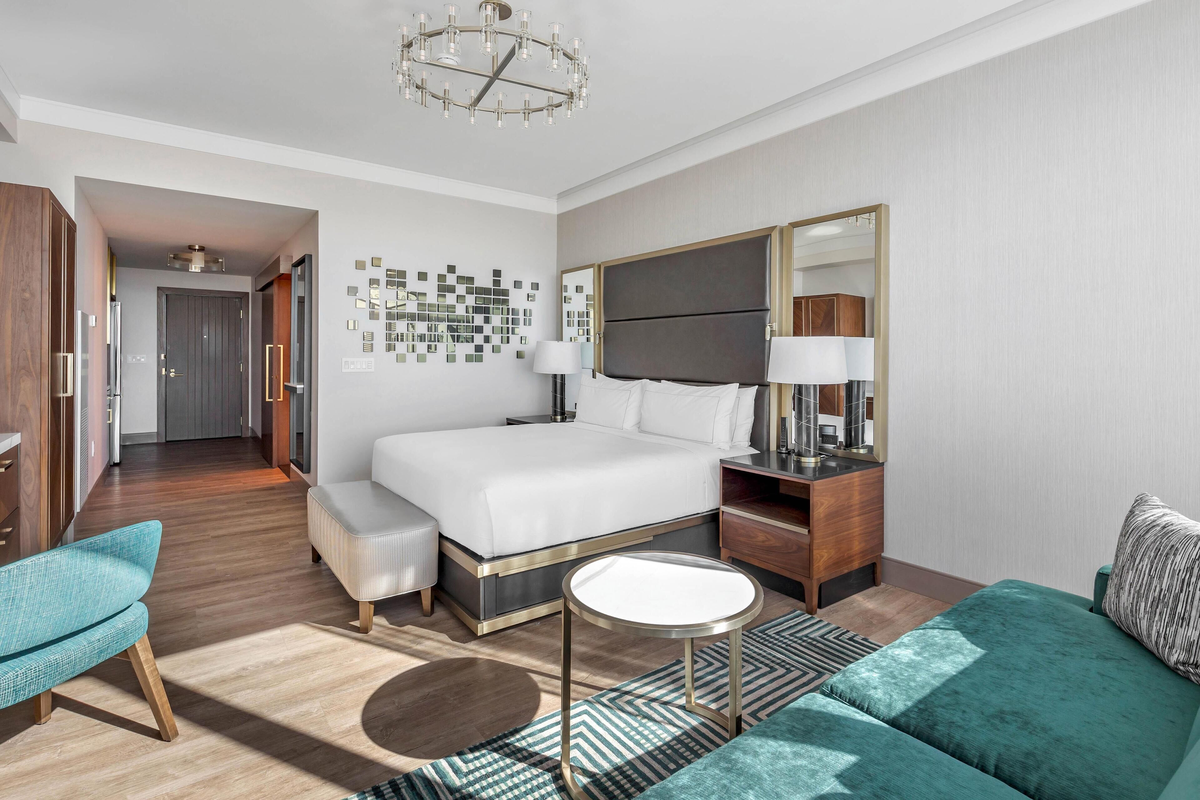 Una habitación de hotel moderna con una cama grande, una zona de estar con un sofá y un sillón, una mesita redonda y un espejo sobre la cama.