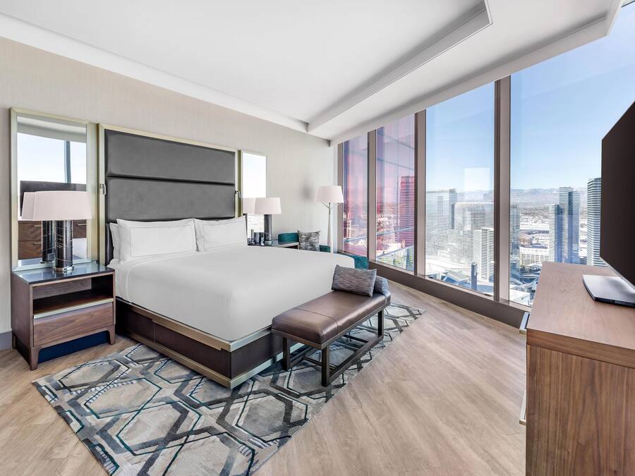 A modern hotel room with a large bed, bedside tables, a bench, and a desk with a laptop.