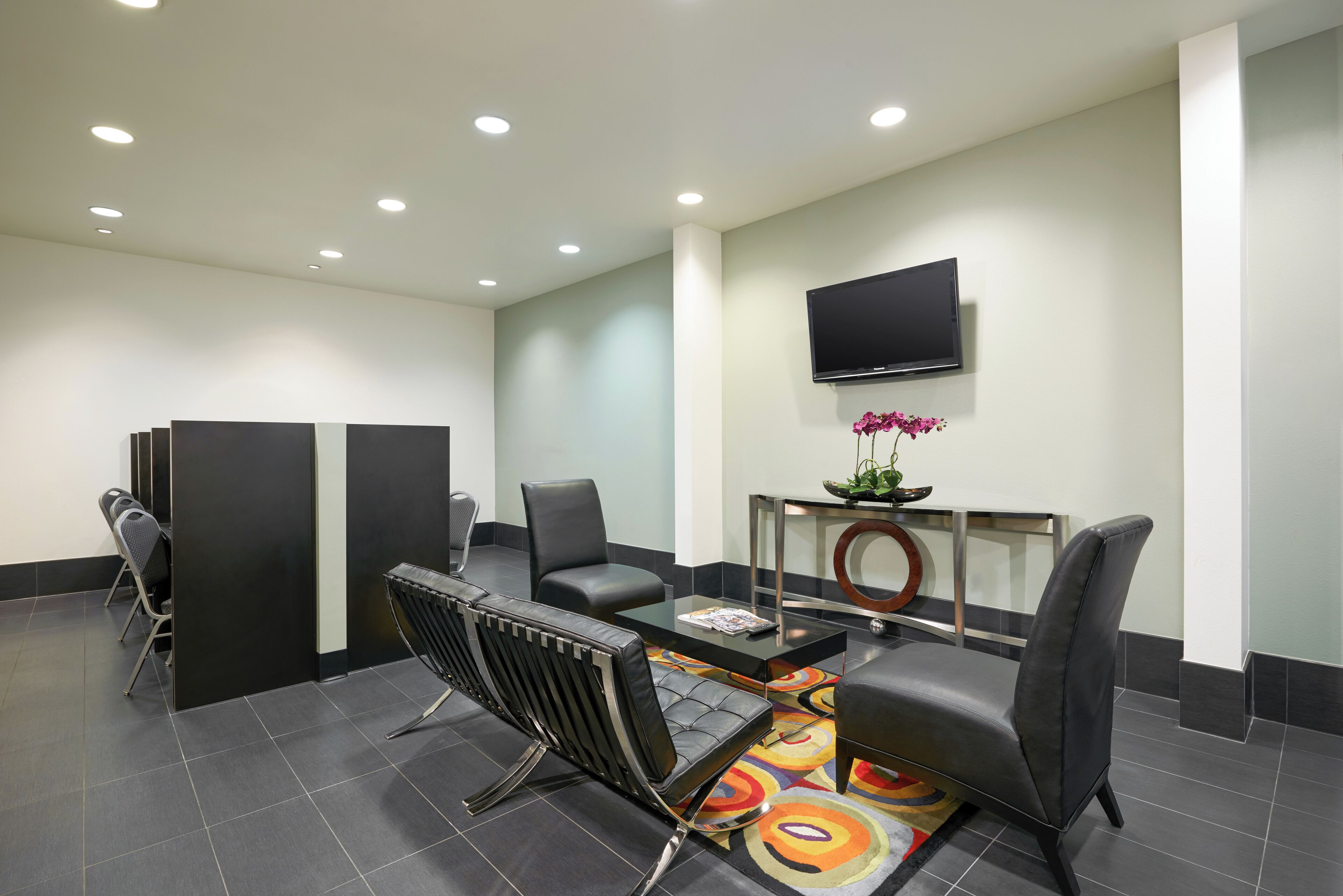 A modern waiting area with a glass table, black leather chairs, a flat-screen TV, and a decorative vase with flowers.
