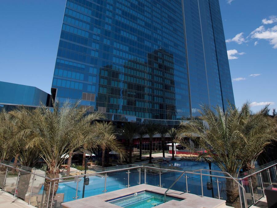 Un hotel de gran altura con fachada de vidrio, una zona de piscina con palmeras y un cielo azul despejado.