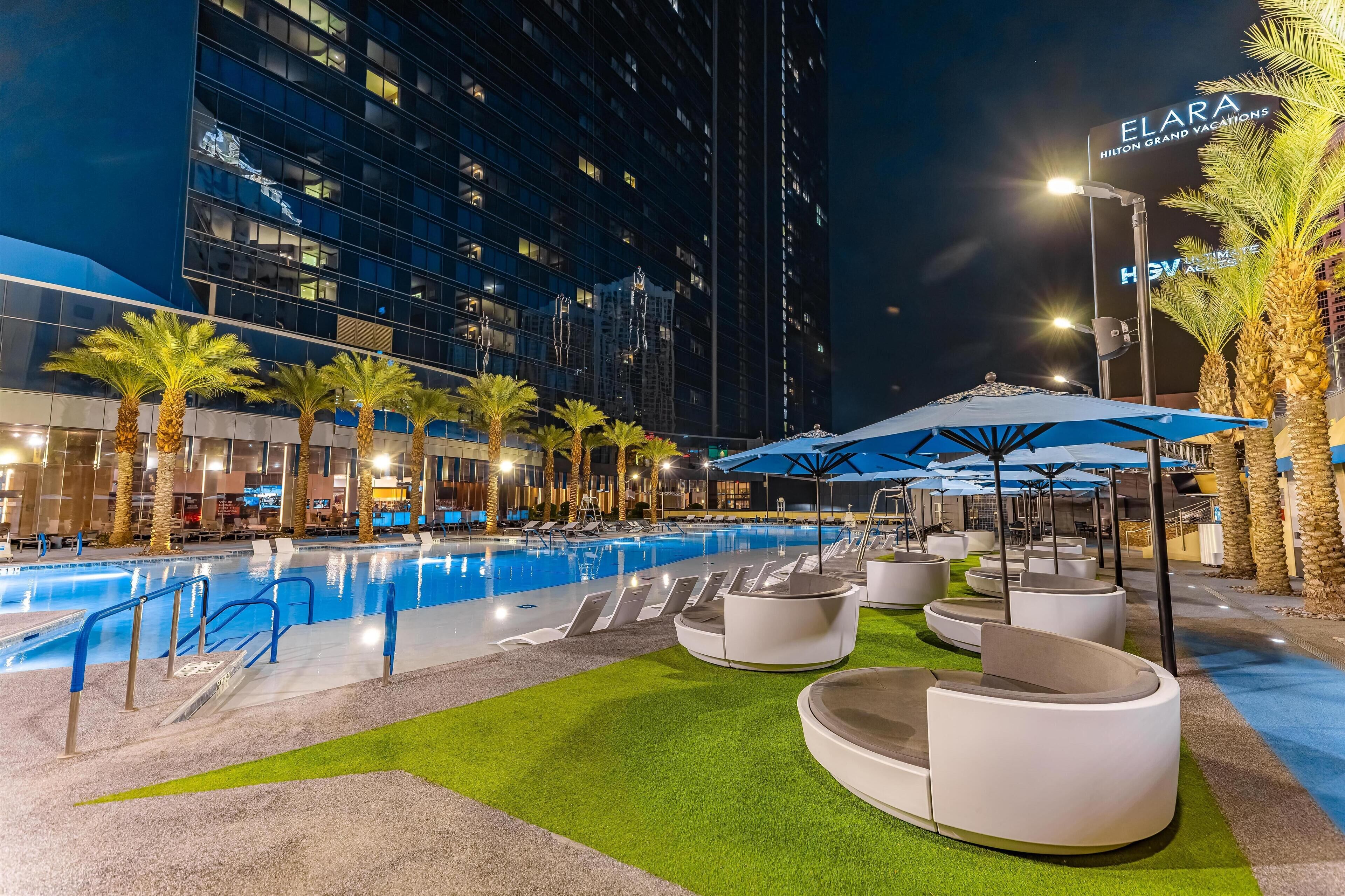 A nighttime pool area with lounge chairs and umbrellas, surrounded by palm trees and modern buildings.