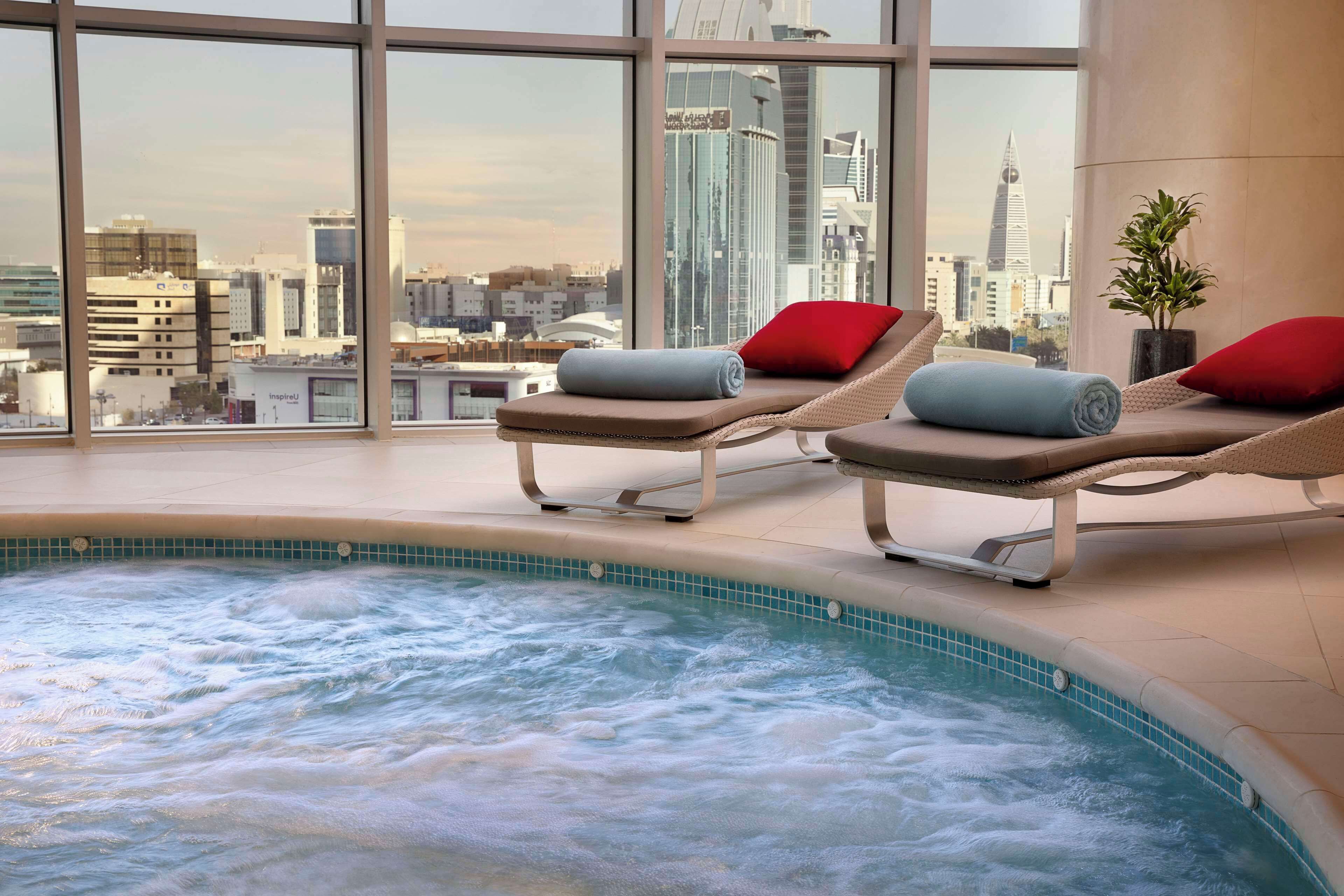 A hot tub area with two lounge chairs, a view of a cityscape through large windows, and a tiled pool edge.