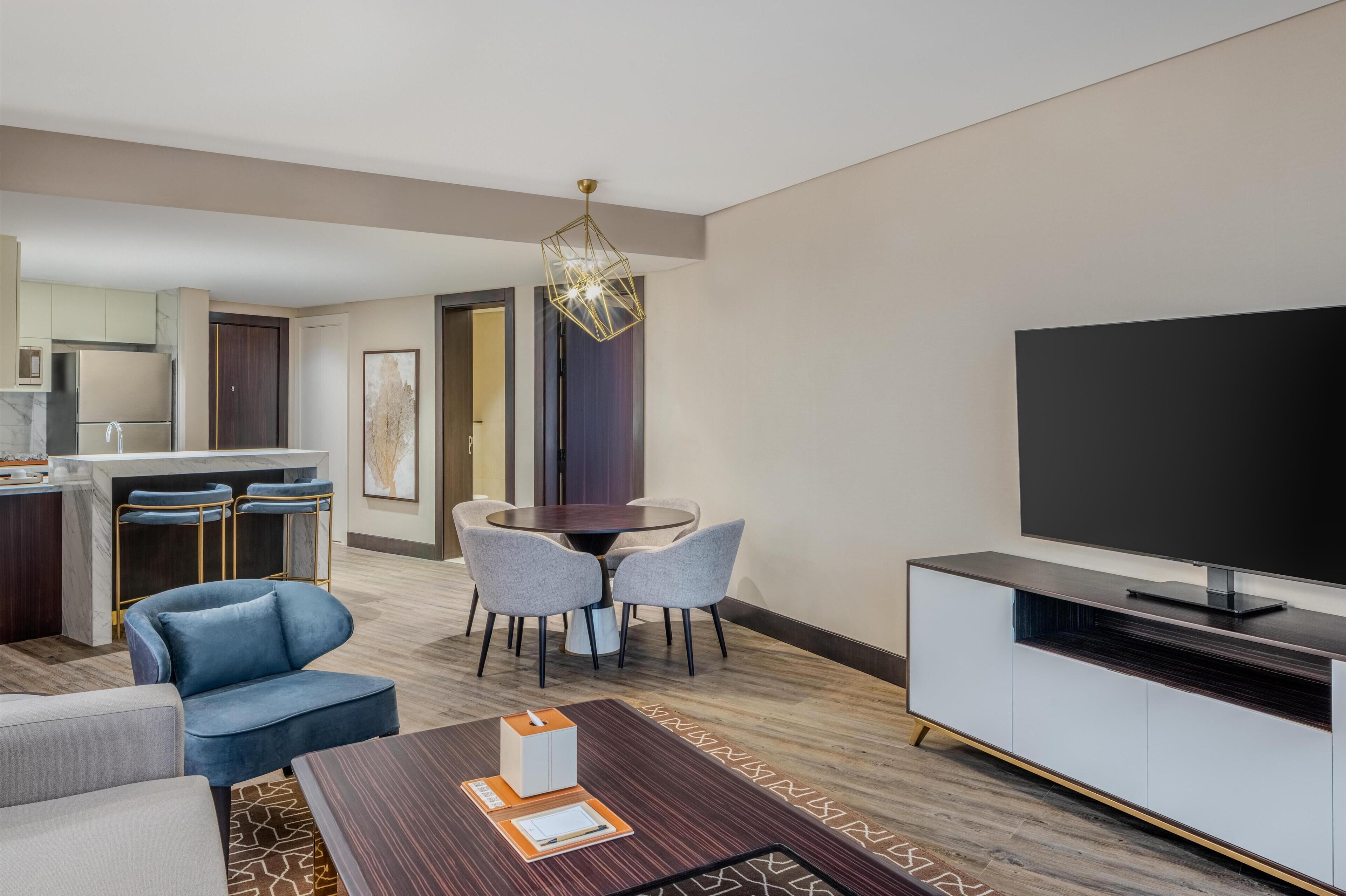 A modern hotel room with a dining area, a sofa, a TV, and a kitchen.