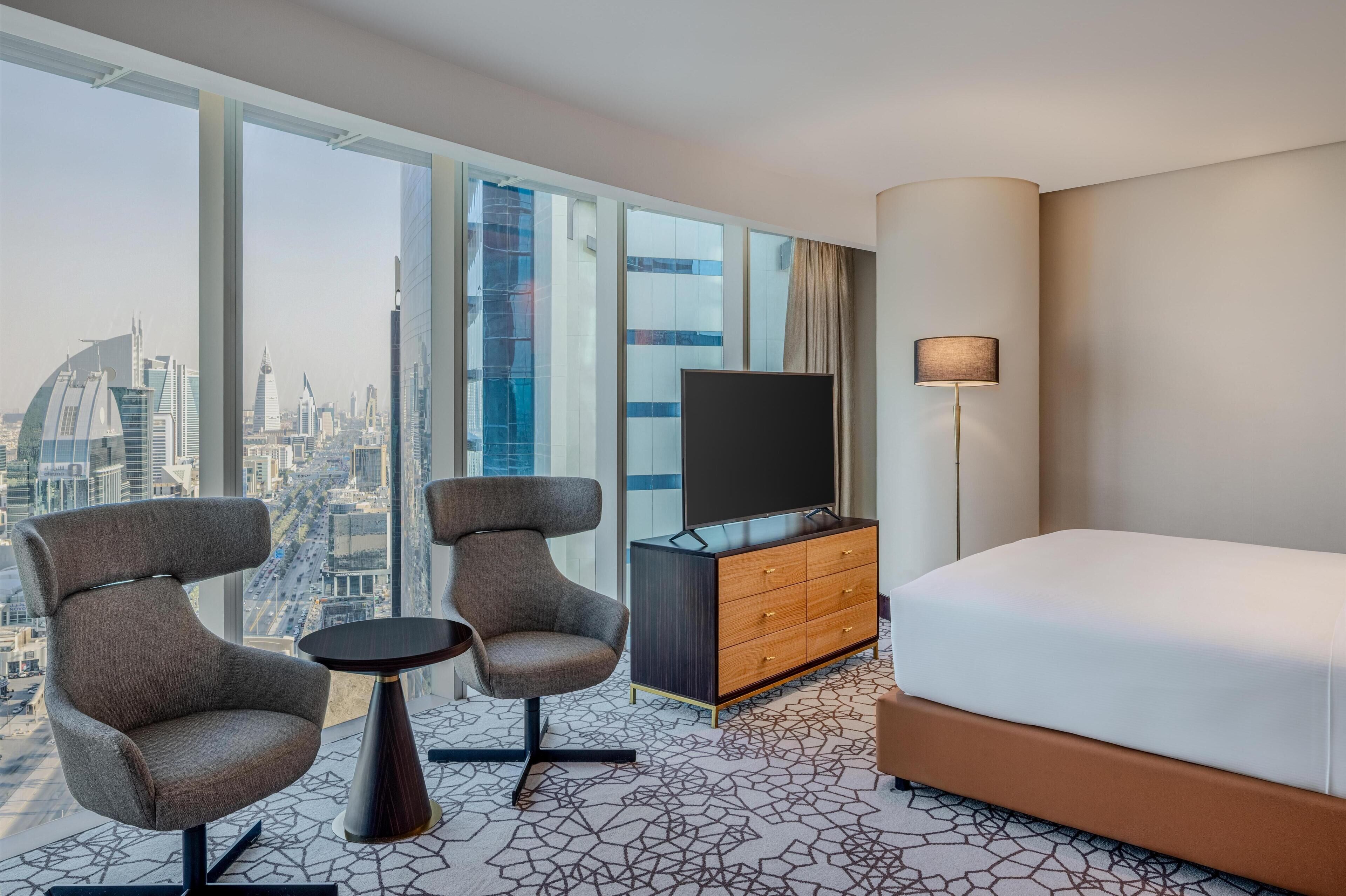 Habitación de hotel con una cama grande, dos sillones, una mesita, un televisor en un mueble y vistas a la ciudad a través de amplias ventanas.