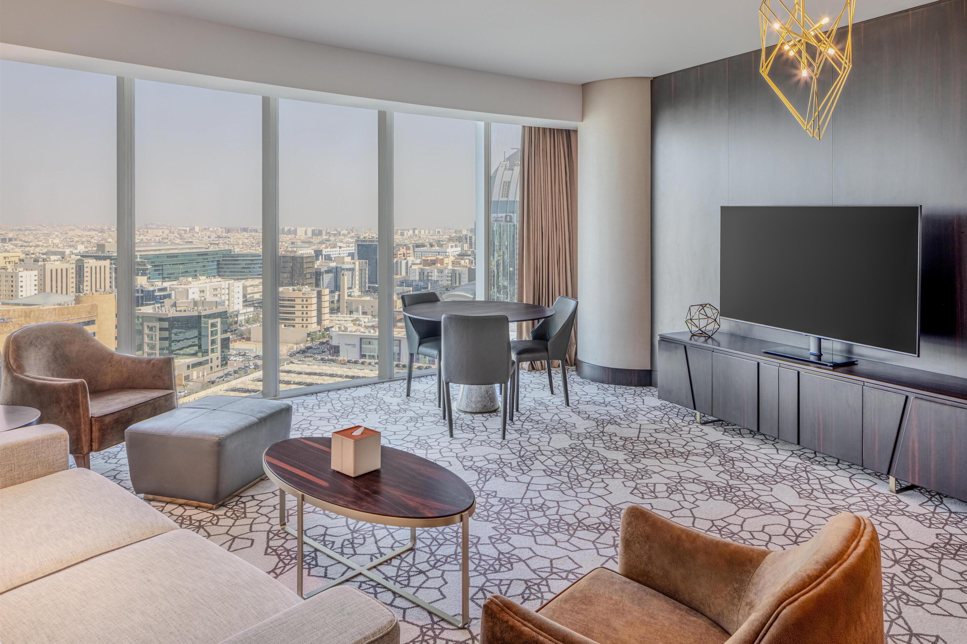A modern hotel room with a large window offering a city view, a flat-screen TV, a dining area, and a cozy seating arrangement.