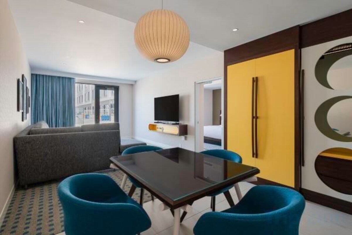 A hotel room with a dining table, blue chairs, a sofa, a TV, and a large window.