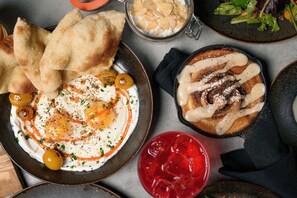 Un assortiment de plats comprenant une assiette avec des œufs, des olives et une sauce rouge, un bol de soupe surmonté d’une crème, et un dessert avec une sauce blanche.