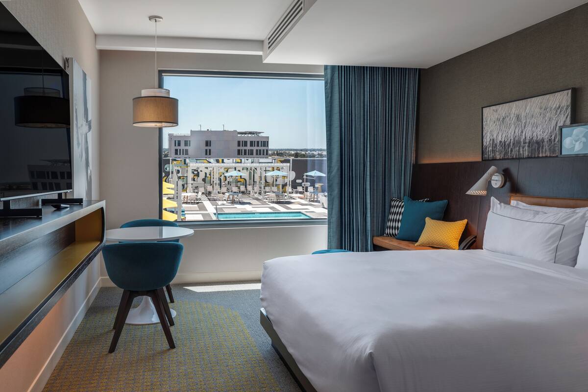 Een hotelkamer met een bed, een bureau, een stoel, een televisie, een raam met uitzicht op een zwembad en gebouwen, en een lampje op het nachtkastje.