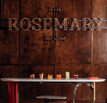 Pared de madera con el texto "THE ROSEMARY ROOM" y una zona de bar con diversas bebidas.