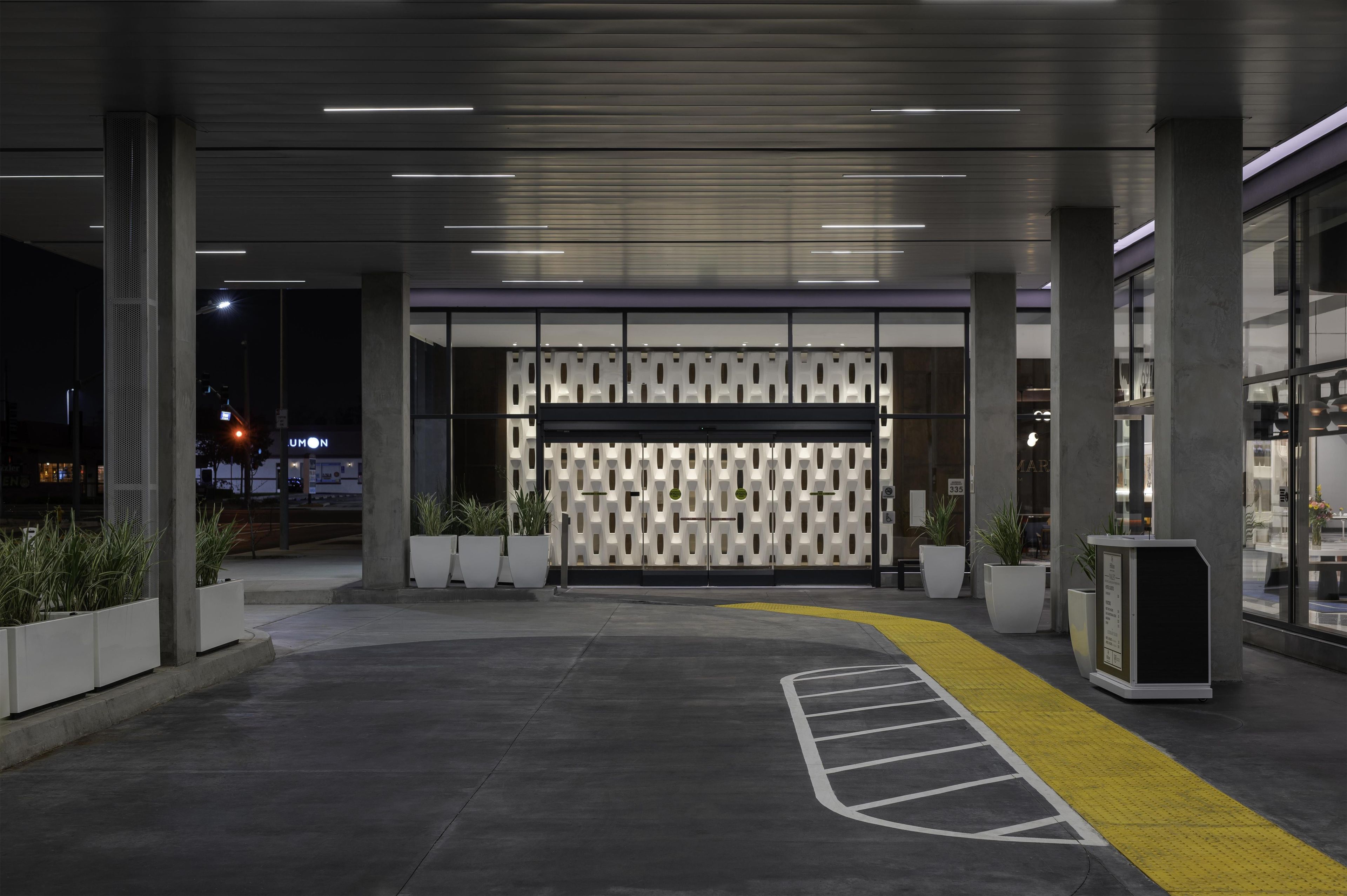 Entrada de un edificio moderno con un muro de diseño, plantas en macetas y un camino pavimentado amarillo con textura.