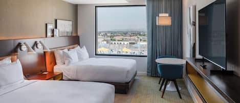 A hotel room with two beds, a TV, a desk, and a view of the city.
