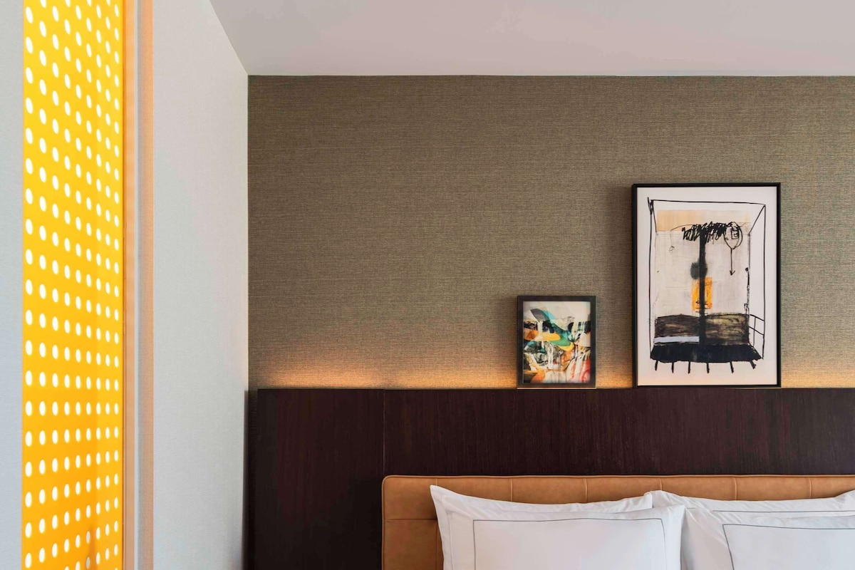 A hotel room with a bed, a nightstand, a wall-mounted artwork, and a window with a patterned blind.