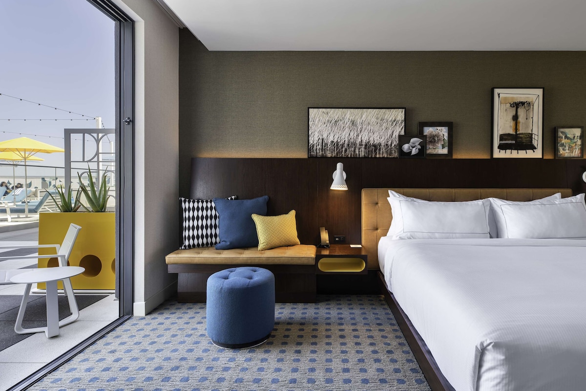 A hotel room with a bed, a seating area with a bench and ottoman, a small table, and a large window offering a view of the outside.
