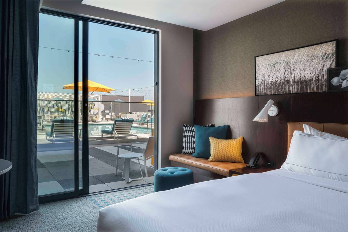 Een hotelkamer met een bed, een bank met kussens, een bureau en uitzicht op een buitenzwembad.