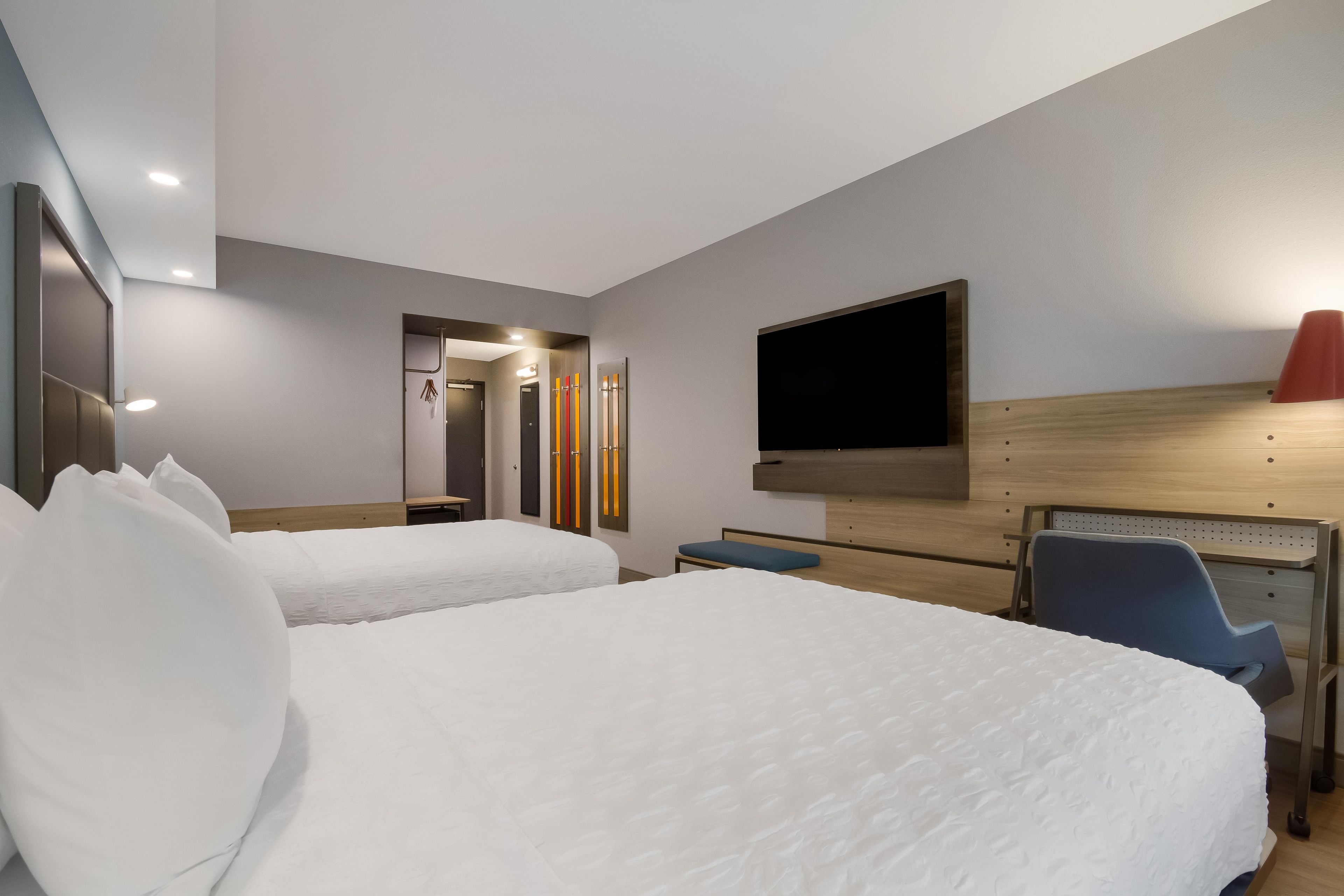 A hotel room with two beds, a desk, a chair, a TV, and a bathroom with a mirror and towel rack.