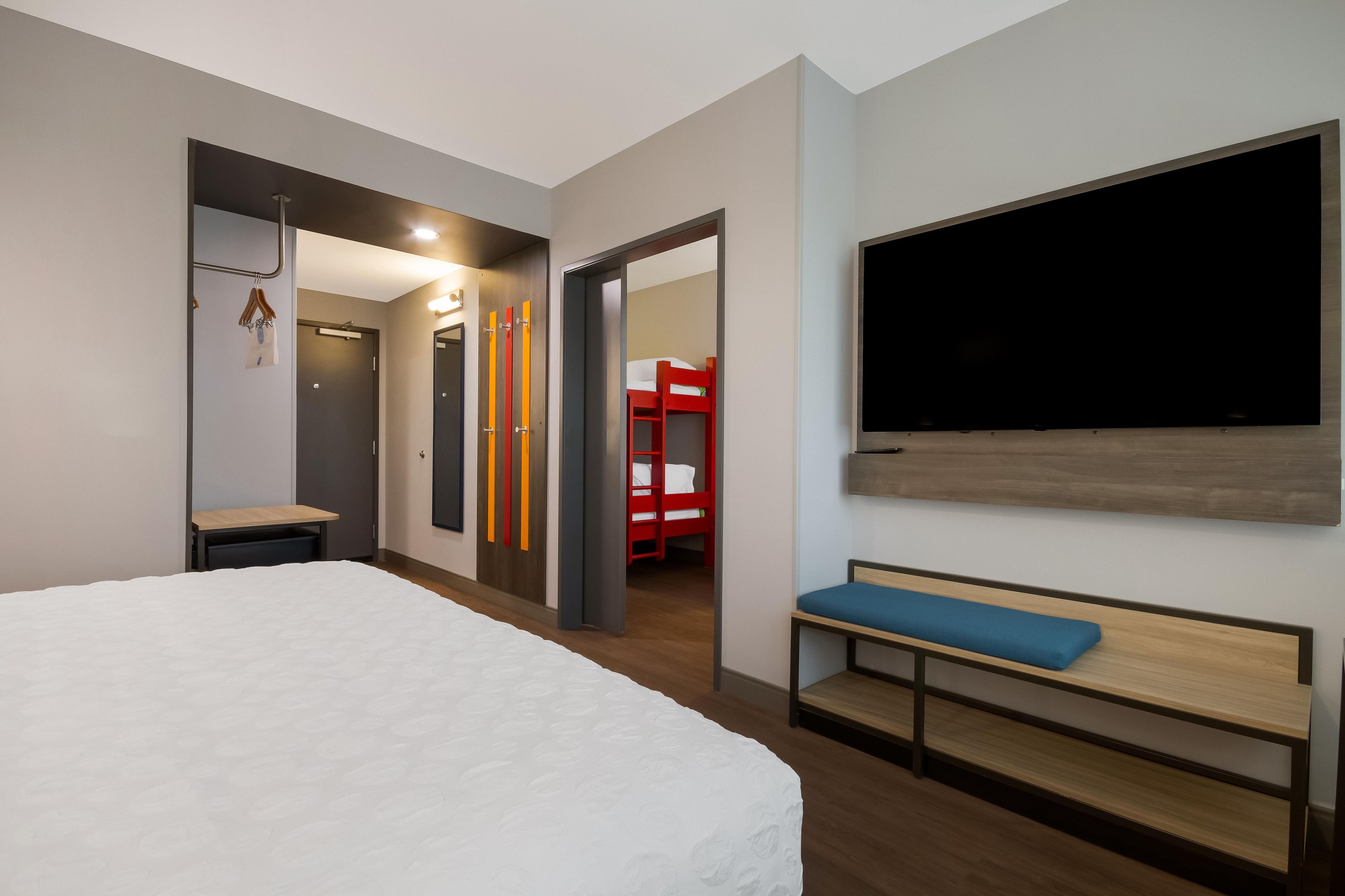 Quarto de hotel com uma cama grande, um banco, uma televisão e uma cama beliche vermelha ao fundo.