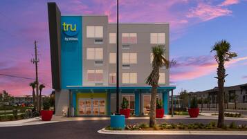A modern hotel with a blue and grey facade, a prominent 'tru by Hilton' sign, and a landscaped entrance area with palm trees and red planters.