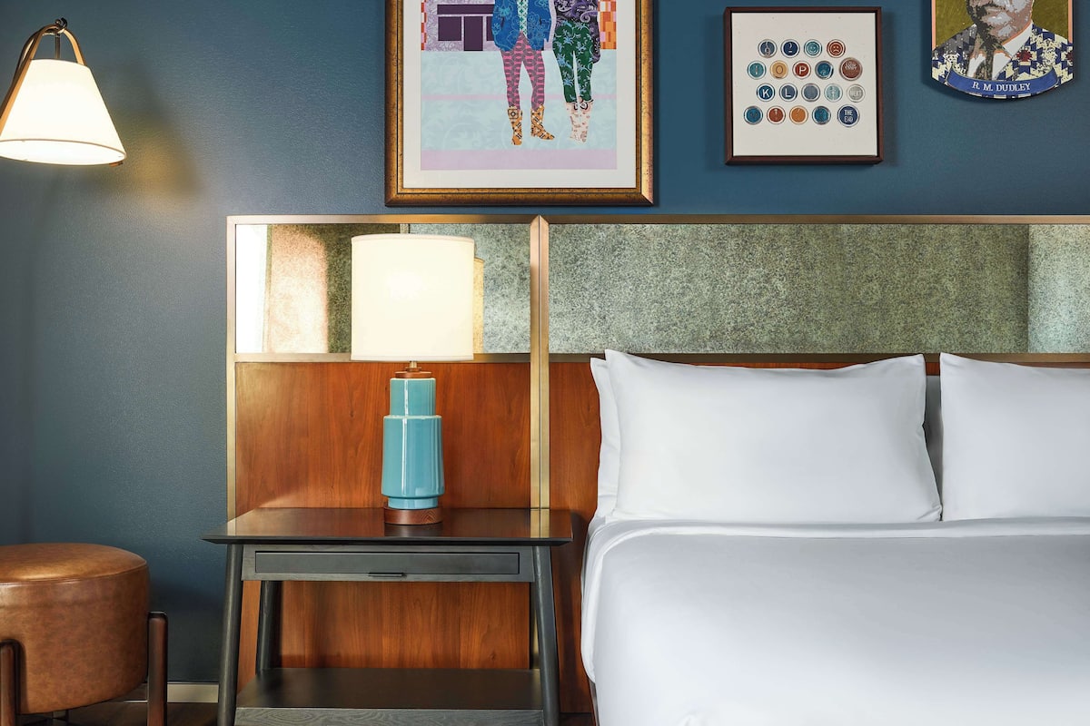 A hotel room with a bed, a nightstand with a lamp, a framed artwork on the wall, and a wooden headboard.