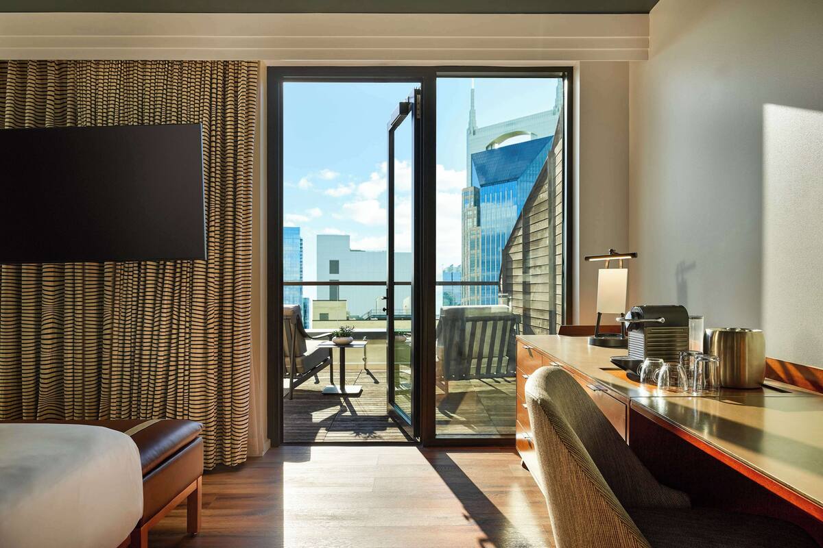 A hotel room with a bed, a desk with a coffee maker, a chair, and a view of a cityscape through a glass door.