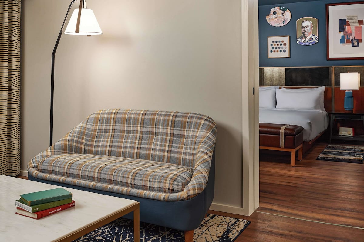 A room with a plaid sofa, a bedside table with books, a lamp, a bed with a headboard, and a wall with framed pictures.